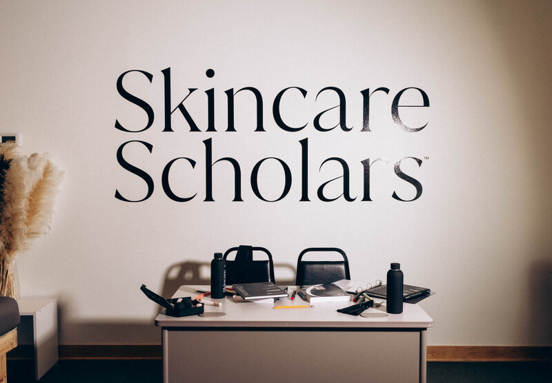 A stylish desk overflowing with skincare books, notes, and tools, with the words "Skincare Scholars" boldly displayed in the background, setting the scene for a day of beauty education and expertise.