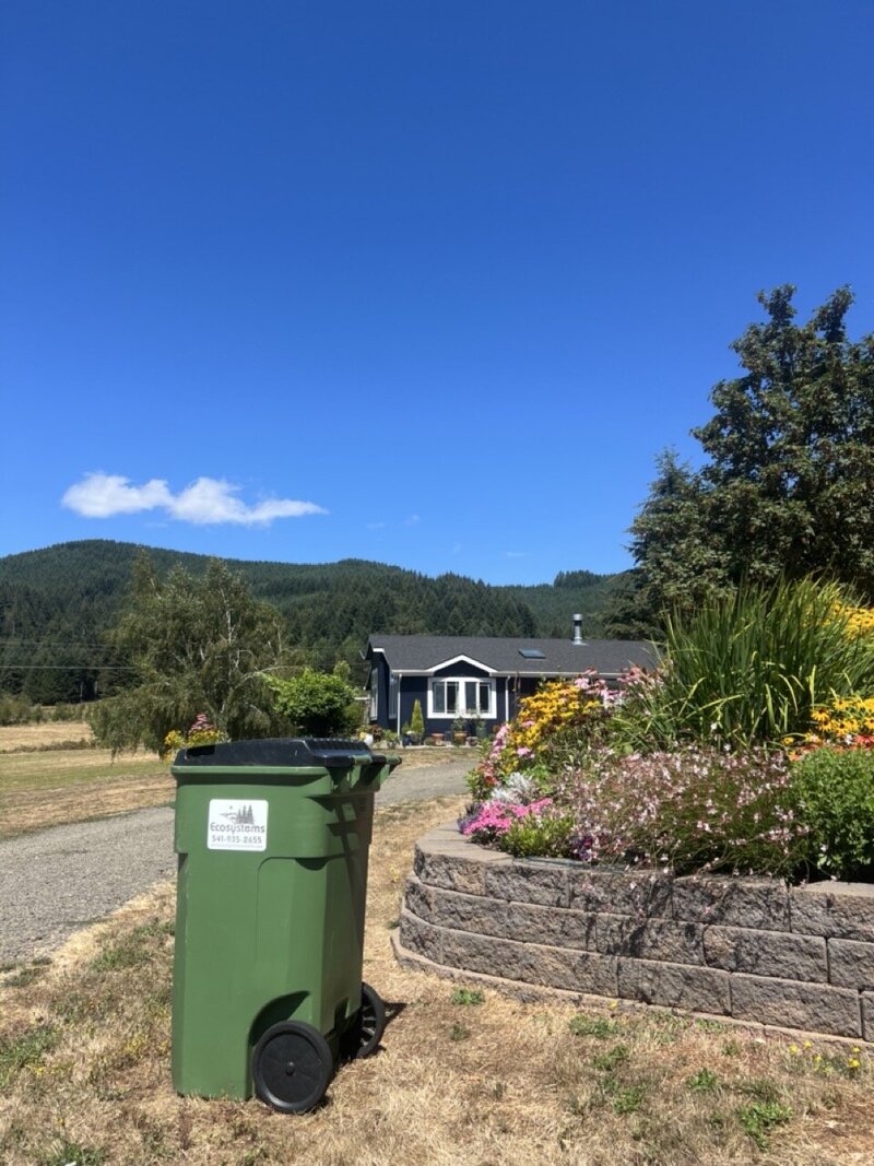 An Ecosystems trash and recycling bin sits at the end of a rural driveway.