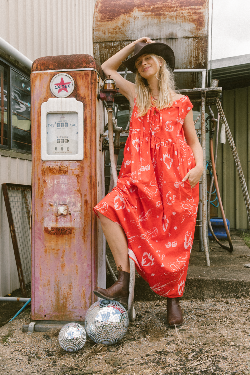 Female model in red dress with cowboy hat and boots on standing next to vintage fuel bowser