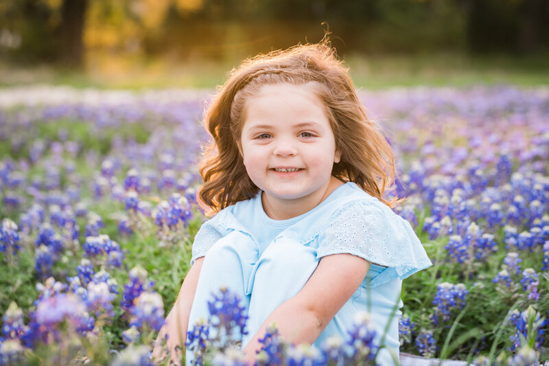 Girl in blue dress sitting in bluebonnet field with sun behind her, Austin Family Photographer