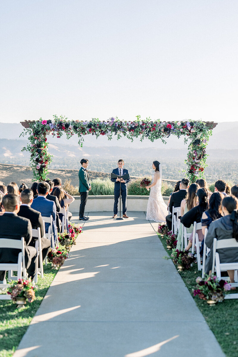 Set against the breathtaking California hills, this couple exchanged vows under a vibrant floral arch that perfectly framed the expansive landscape. With romantic hues of purple, pink, and green, the ceremony’s elegant decor complemented the natural beauty of the venue. Surrounded by family and friends, the couple shared a heartfelt moment that captured the essence of a classic California wedding. The soft sunlight and panoramic views enhanced the magic of this outdoor celebration, creating an unforgettable experience.