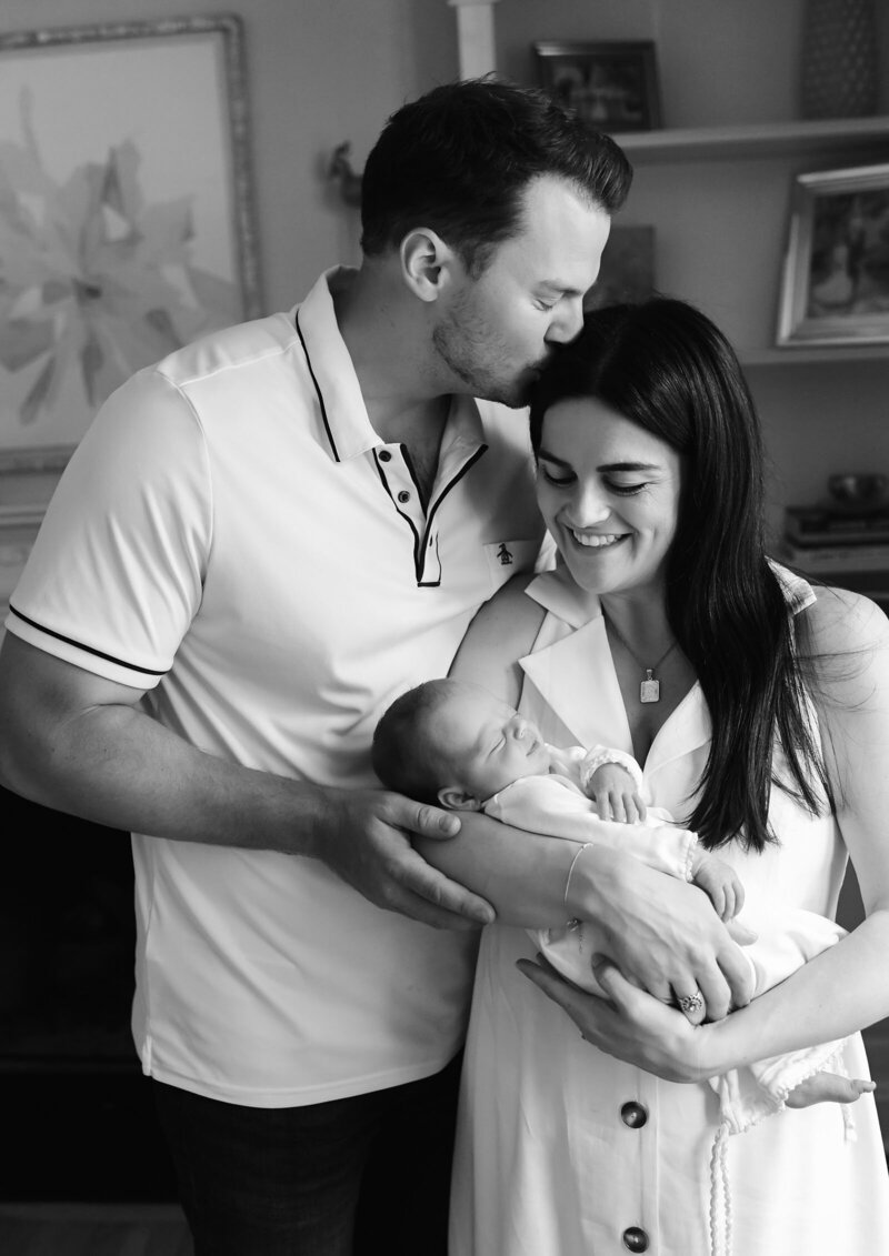 A black-and-white photo by a St. Louis photographer shows a couple with a newborn baby. The man, wearing a polo shirt, kisses the woman's forehead. The woman, in a sleeveless dress, cradles the sleeping baby in her arms. They stand in a cozy room with shelves and framed pictures in the background.