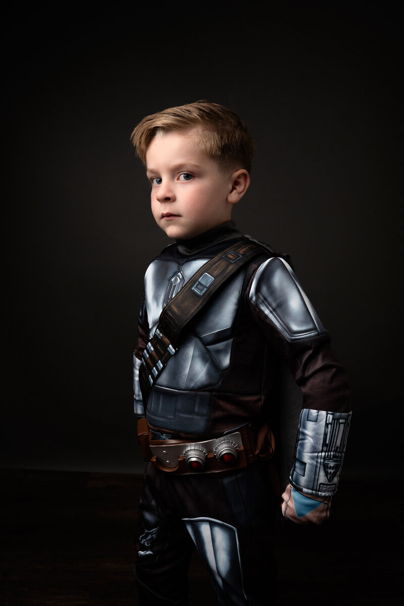 Young boy in Mandalorian Costume unmasked with closed fist in front of black backdrop with dramatic lighting.