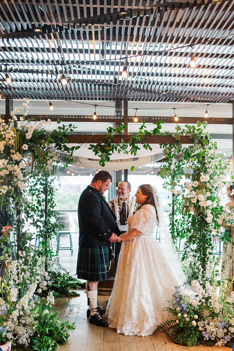 Scottish groom with jewish bride holding hands and looking at one another under a wooden chuppah adorned with greenery and white flowers with a rabbi inside District Winery ballroom 