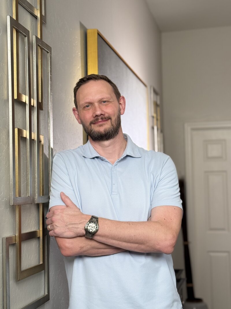 man smiling and leaning against a wall indoors in a light blue polo shirt with his arms crossed displaying a casual, kind, confidence.