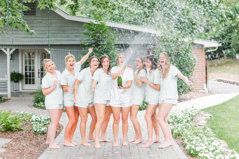 A bride wearing white pajamas and her bridesmaids wearing sage green pajamas crack up laughing as they pop a bottle of champagne