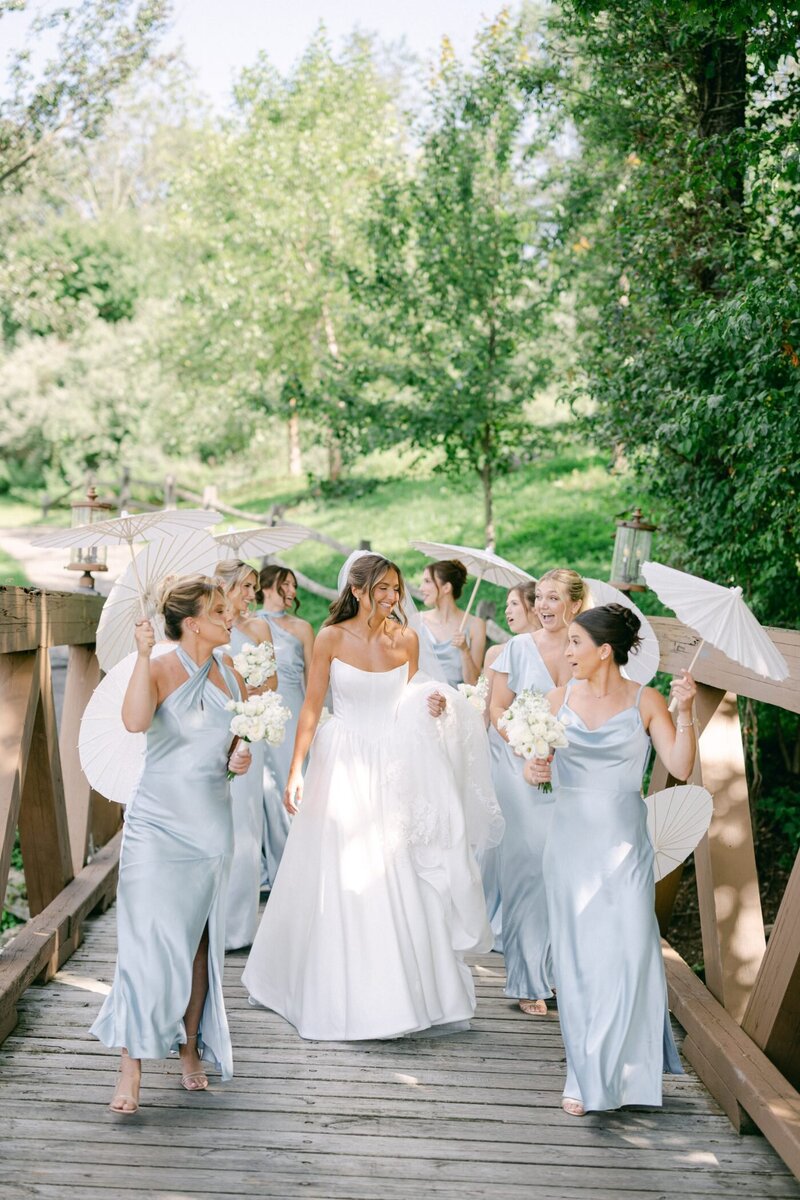 Anya and her bridesmaids crossing the wooden footbridge with white parasols