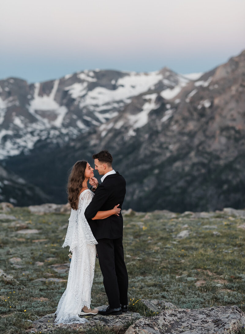 A groom lifts his bride's chin and they are smiling at each other. there is a mountain behind them