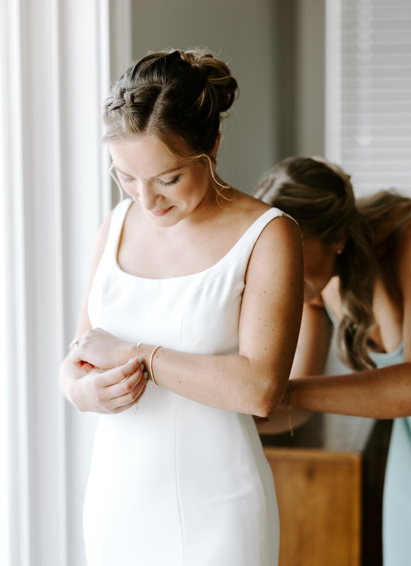 Bride plays with her bracelet while bridesmaid buttons up her wedding dress in Cape Cod, Massachusetts