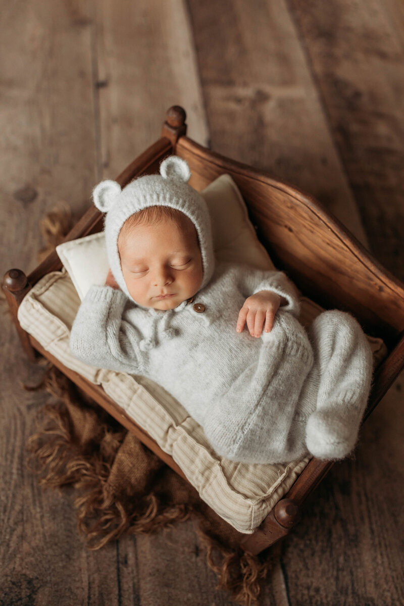 newborn baby in a teddy bear outfit laying on a tiny wooden bed