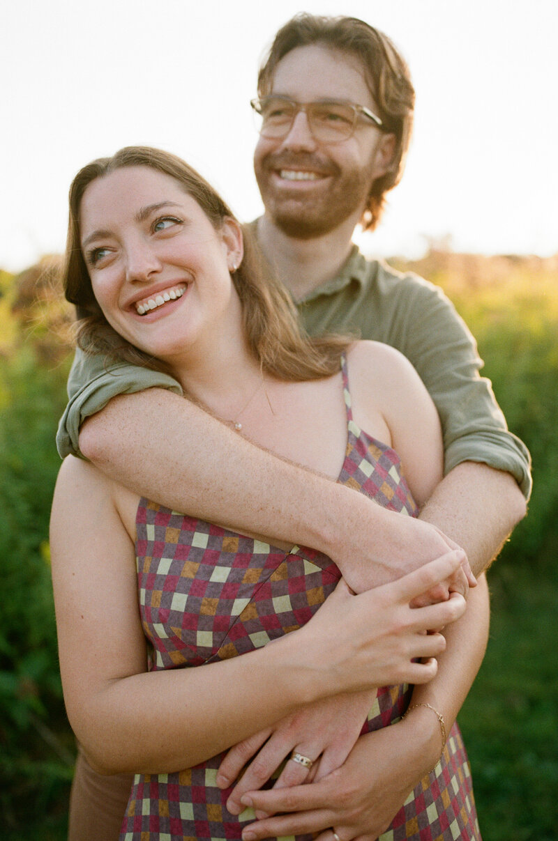 A man and woman share a warm embrace in a sunlit field, surrounded by tall grass and blooming wildflowers.