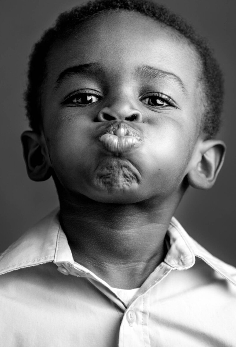 A young child in a buttoned shirt makes a playful, exaggerated pout against a plain background. The monochrome image conveys a lighthearted, humorous tone.
