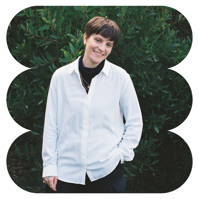 Hayley Kendall | Owner of Tidy Books Club. ID: Woman  with short brown hair wearing a white button down shirt smiling for the camera and standing in front of a deep green bush.