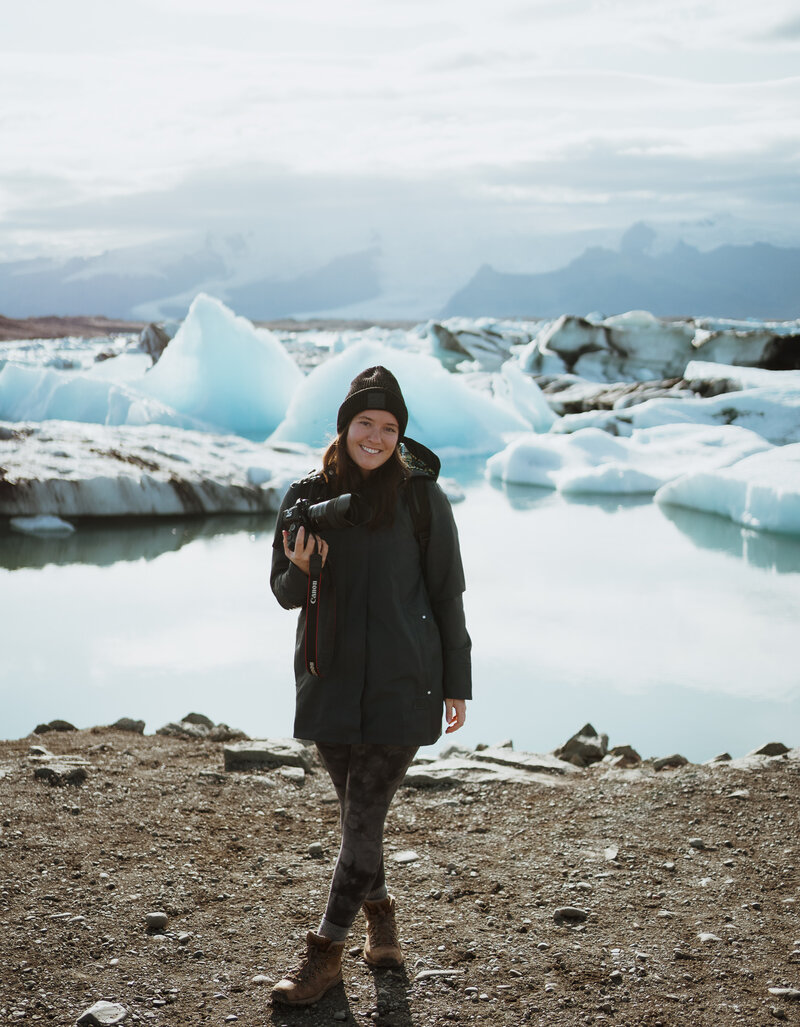 woman smiling at the camera. She is standing in front of ice bergs and a glacier lake. she is wearing a black beanie and a long navy rain jacket. she has on black leggings and hiking boots. It is sunny outside and there are blue tints behind her.