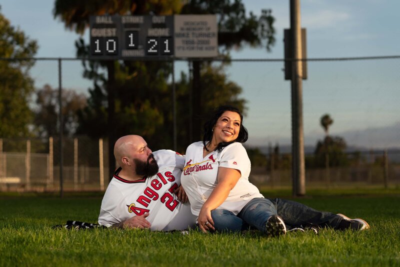 Man laying on outfield grass with woman leaning on him being silly. Scoreboard in background has their wedding date.