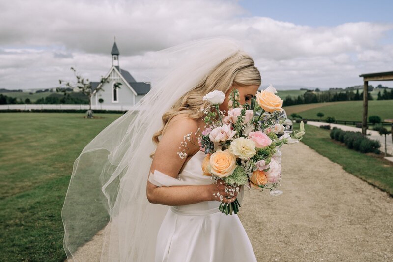 candid shot of bride with pastel bouquet in front of wooden church at farm chapel waimumu