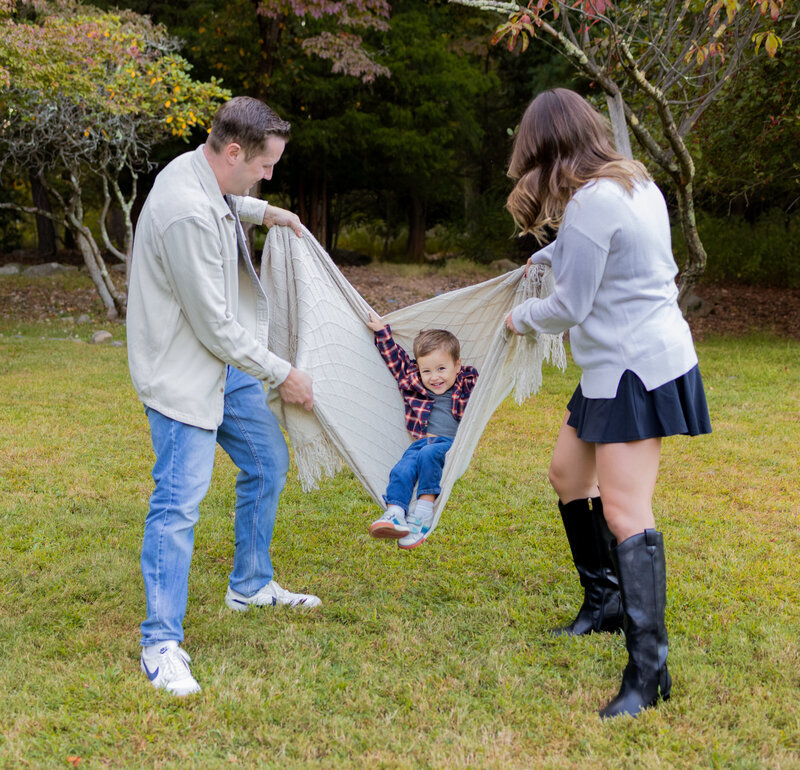 Mom and dad swinging their toddler in a blanket while he laughs during an outdoor photo session near Mahwah, NJ.