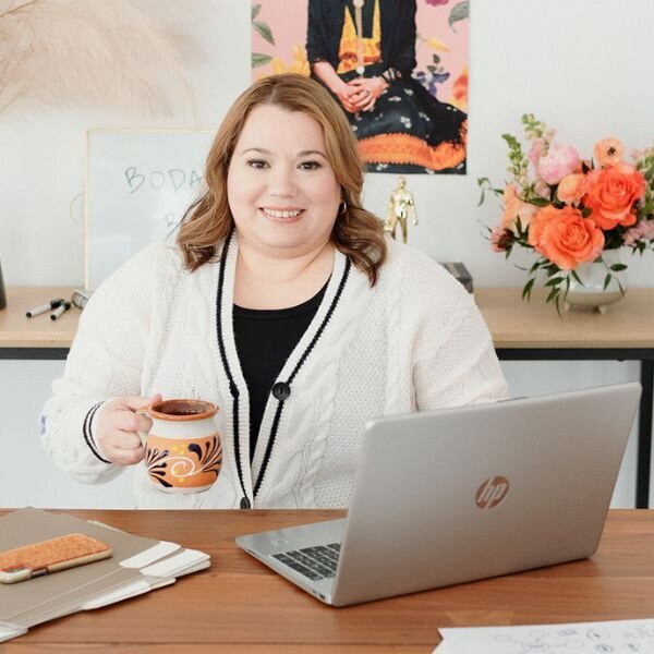 A woman with light brown hair, wearing a white cardigan over a black top, sits at a desk with a laptop and a mug in hand. The background features a shelf with flowers and framed artwork, adding a warm and inviting feel to the workspace.