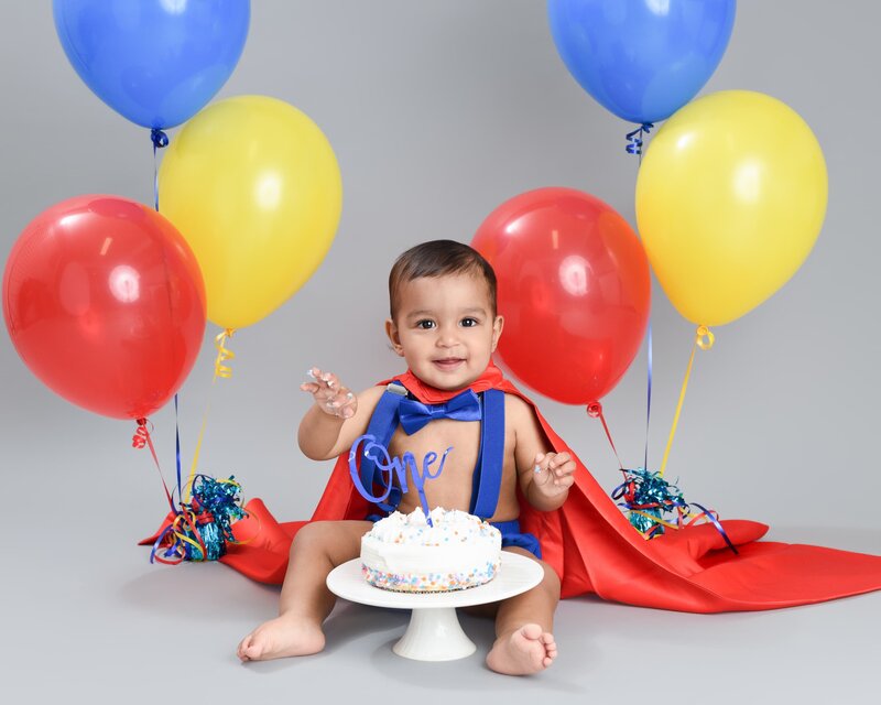 A baby boy dressed as a superhero in a red cape and blue outfit, sitting with a cake and surrounded by red, yellow, and blue balloons.