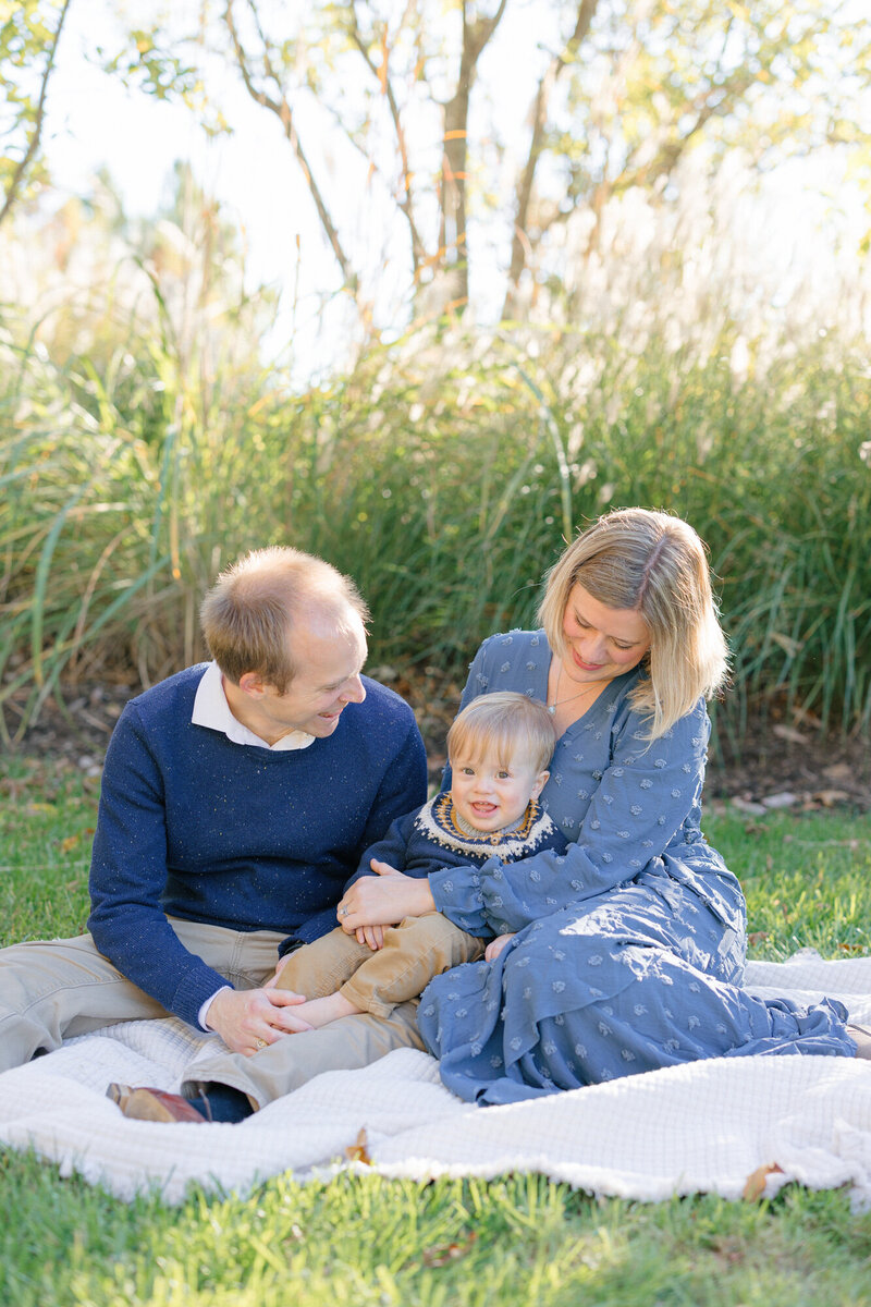 Family of three snuggling on a white quilt in a green park during their family photography session in louisville kentucky