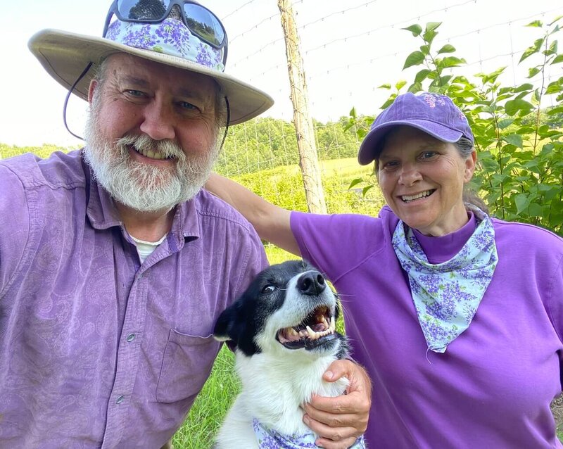 Owners Wil and Rebecca Rivers smiling on farm in purple shirts and hats with dog, Delilah 