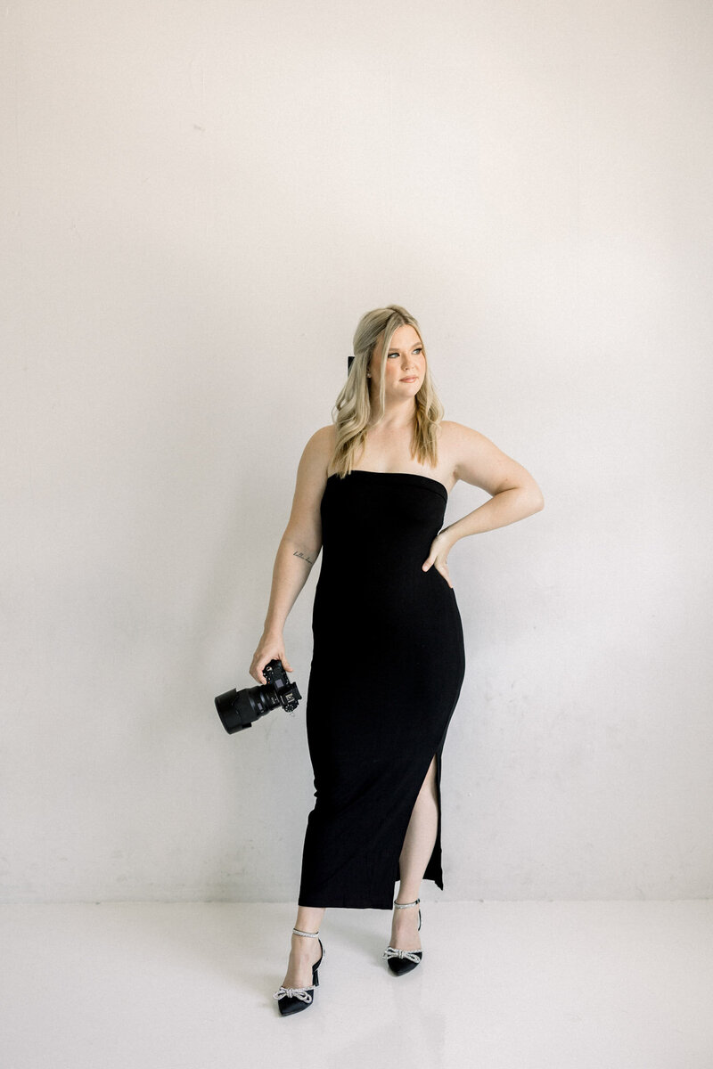 Full-length portrait of a Scottsdale photographer in a black formal dress holding a camera, standing against a light neutral background.