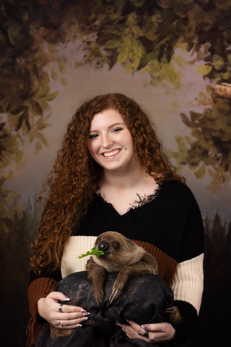 girl-with-red-hair-holding-sloth-on-pillow-in-studio-arlington-tx-with-forest-background