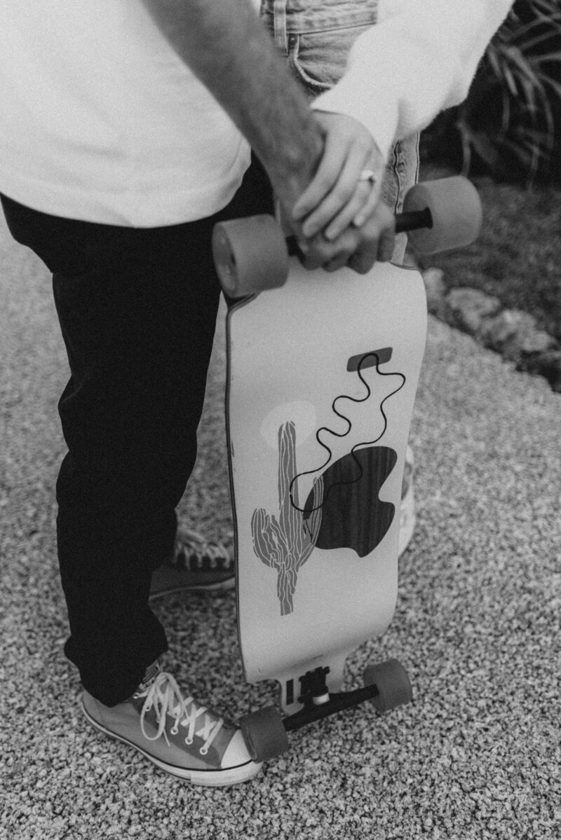 a longboard with a saguaro on the back of it while dom's hand rests on her husbands who's holding the top of truck on the board, picture is in black n white with grain