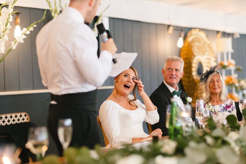 A woman in a white wedding dress smiling and wiping a tear from her eye sits at the top table, listening to a her husbands speech. Another man and a woman sit beside her, surrounded by greenery and glasses.