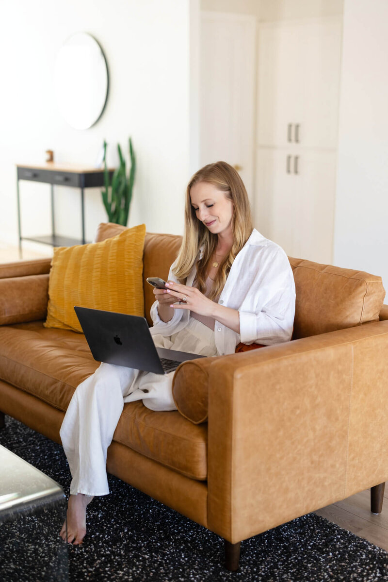 Mary Pernicano, San Diego realtor, sitting on a modern tan sofa while working on her laptop and phone during a client consultation — Mary Pernicano Real Estate.