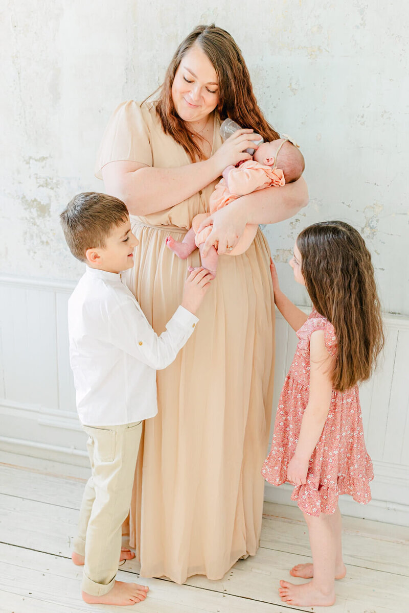 Mom gives her baby a bottle while her two older kids look on at a newborn session by a Boston Newborn Photographer