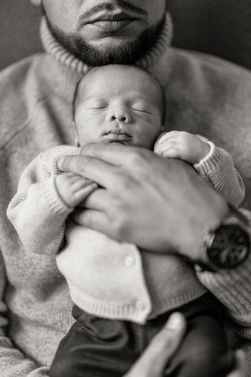 Newborn baby sleeping in a parent's arms, captured in a soft black and white portrait.