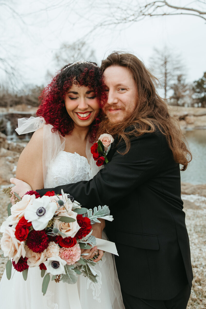 A newlywed couple shares a tender moment, with the bride kissing the smiling groom on the cheek, seated on patterned tiles amidst lush greenery