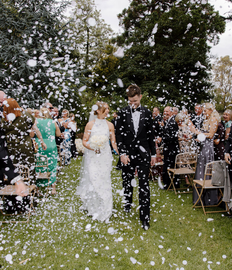 A vibrant and joyful moment captured at Burnley Gardens in Richmond, showing the bride and groom walking hand in hand down the aisle after their wedding ceremony, surrounded by a shower of white flower petals. The bride is wearing a sleek, elegant halter-neck white gown, holding a bouquet of white flowers, while the groom looks dapper in a classic black tuxedo with a bow tie. The couple smiles softly as they walk, while guests, dressed in colorful formal attire, clap and throw petals in celebration. The lush garden setting features tall trees, greenery, and wicker chairs, adding to the romantic and natural ambiance of the outdoor ceremony. The flying petals create a dynamic, festive atmosphere, adding a sense of movement and excitement to this beautiful moment before the couple heads to their reception at Cutler and Co.