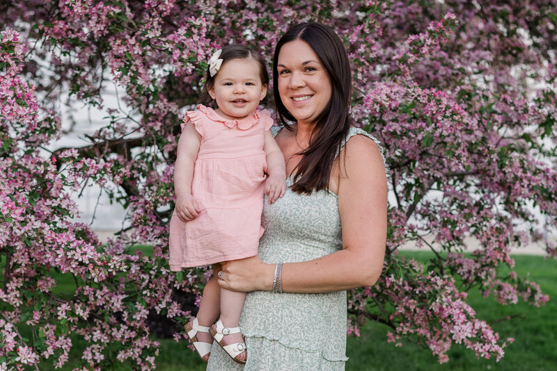Mother and daughter smiling at the camera during a mommy and me photoshoot in front of blossom trees.