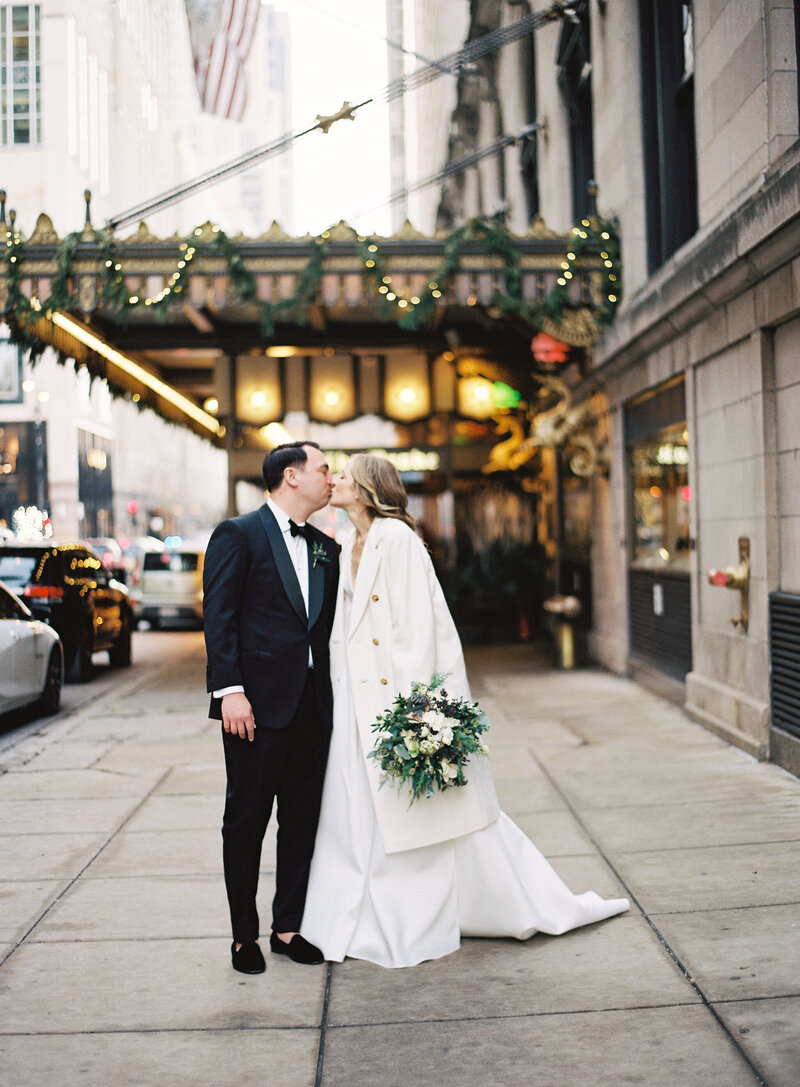 Bride and groom share a kiss on a city street, with the bride in a long white gown and a fur coat, holding a large green and white floral bouquet. Festive garlands and lights adorn the building facade behind them, adding a romantic winter holiday atmosphere to the scene.