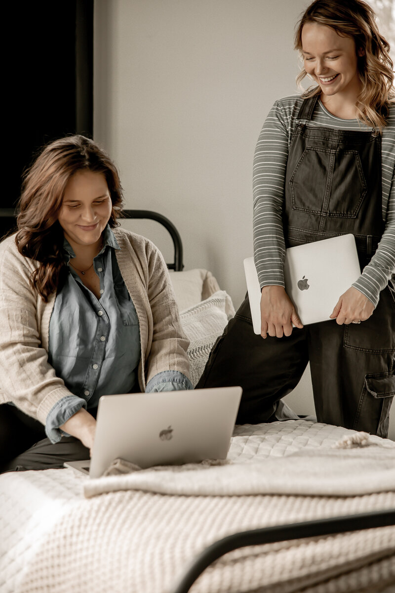 Lacey and Amber, co-founders of First & Third Designs, smiling while Lacey sits on the bed working on a laptop and Amber kneeling on the bed looking over her work