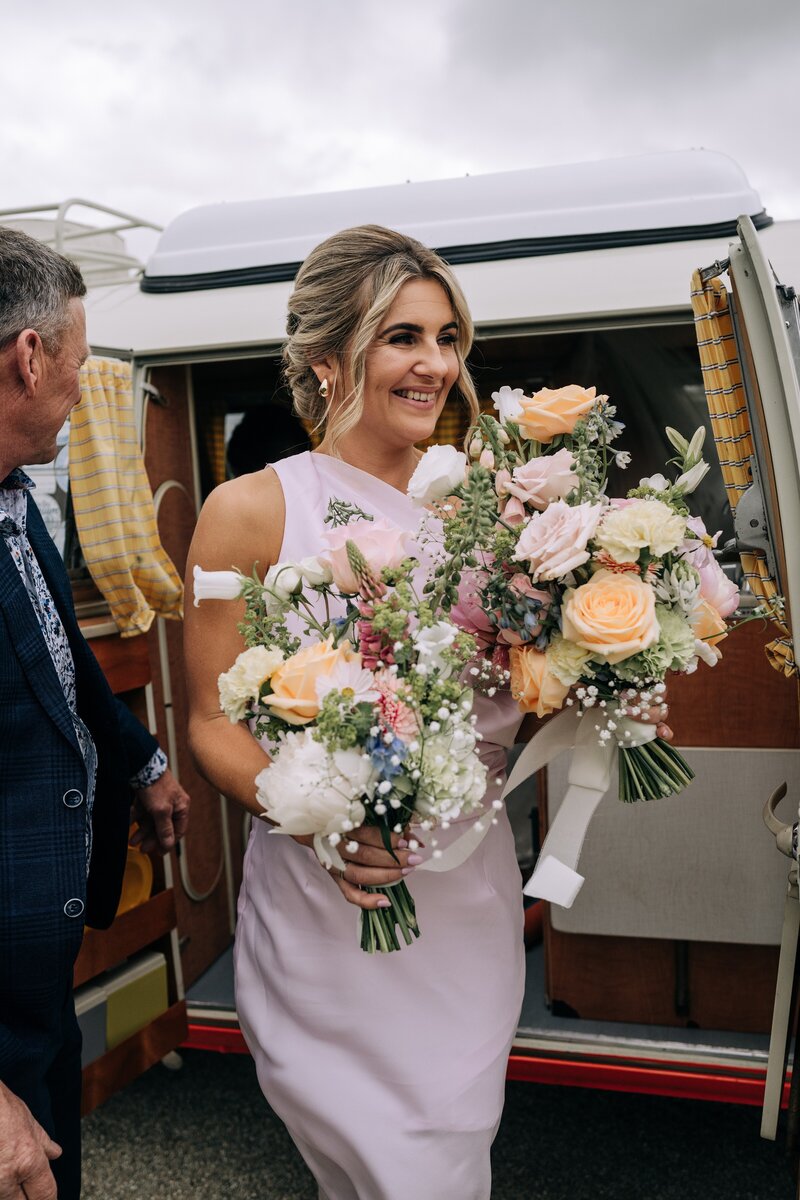 bridesmaid in pink with pastel bouquets gets out of kombi van at farm chapel wedding near gore
