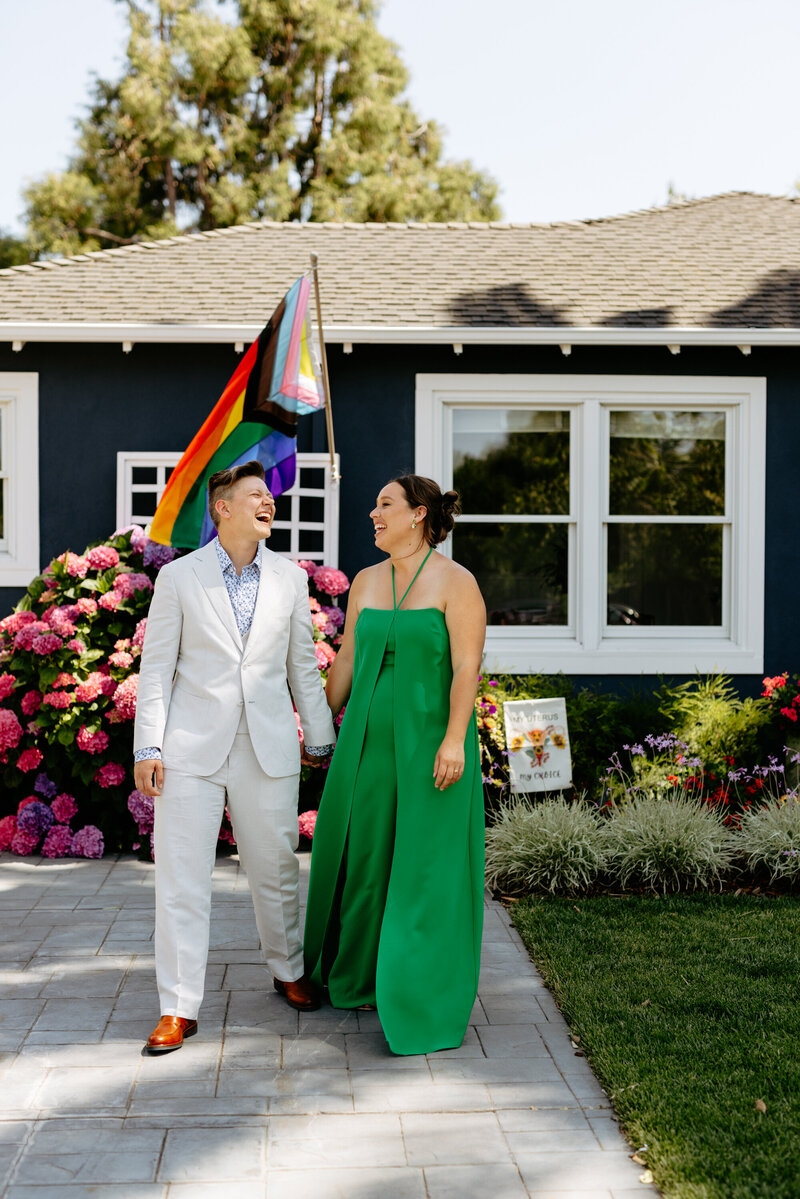 lgbtq couple laughing walking together with pride flag flowing behind them