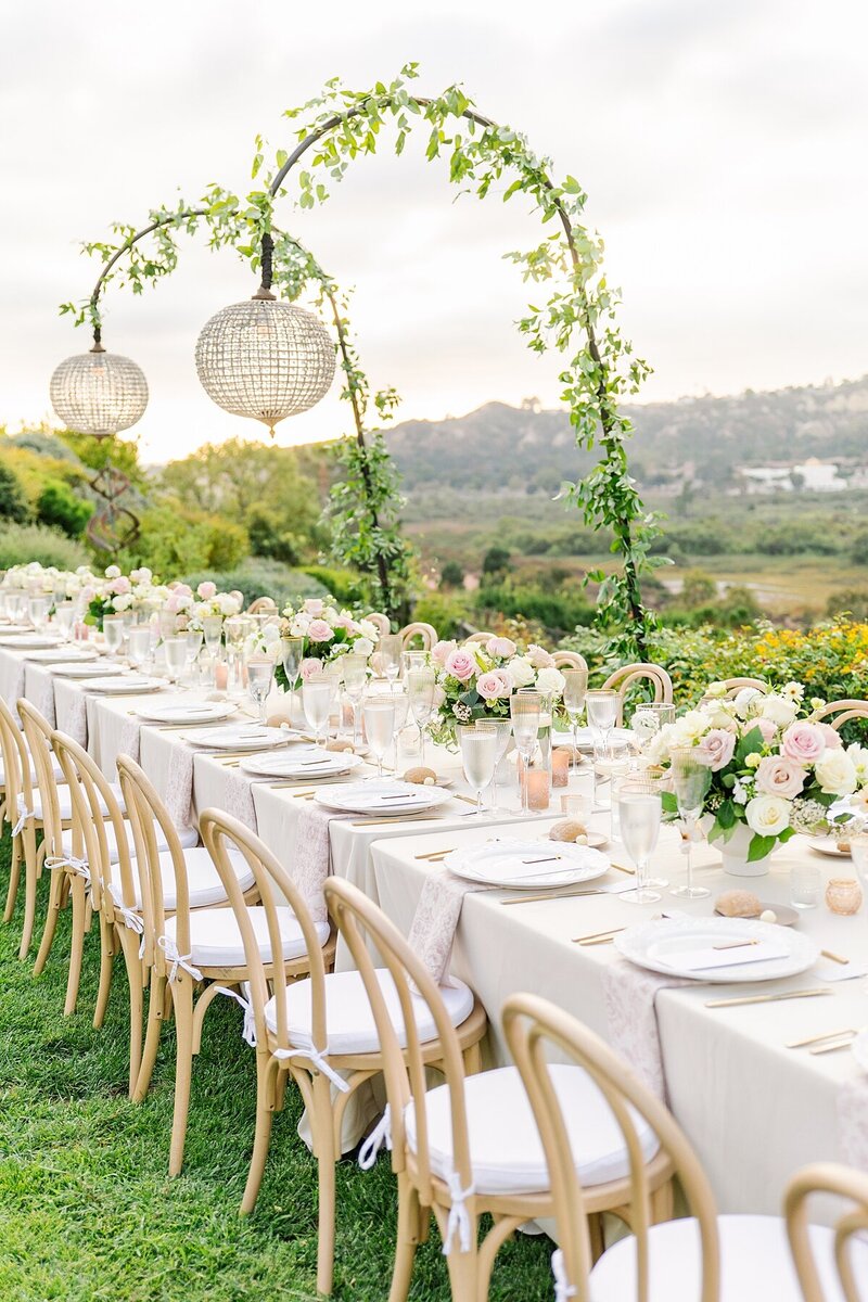 Wedding reception at a private residence in Rancho Santa Fe, California. with tan linens and blush napkins and floral centerpieces. Wooden chairs with white seat covers are pulled up to the long tables. Ivy-covered crystal lanterns hang overhead the tables as the sunset glows behind it all.