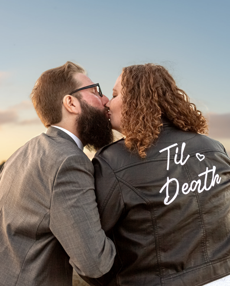 bride and groom kiss in front of a monterey sunset. The Bride wears a leather jacket that has the words Til Death embroidered on it.