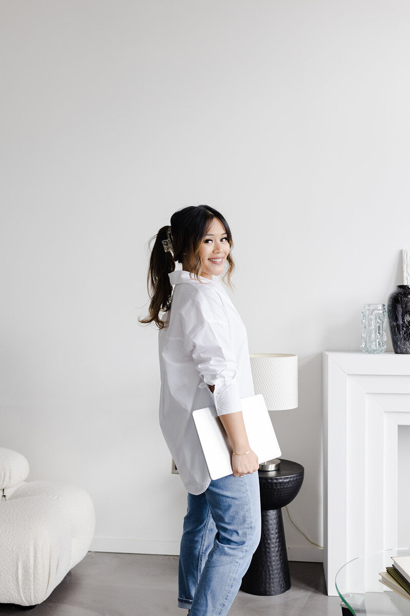 woman business owner in blue jeans and a white button down shirt standing and smiling at the camera with her laptop in hand