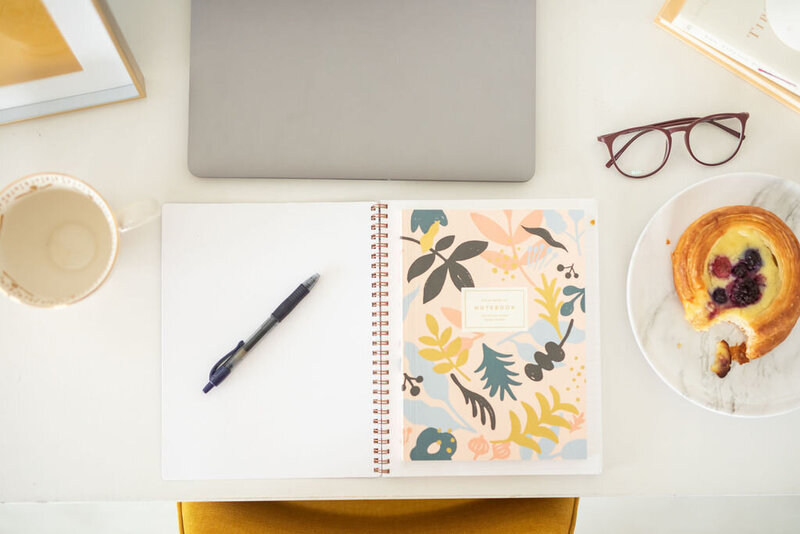 The desk of an email copywriter includes notebook, pen, croissant, glasses, laptop, and mug