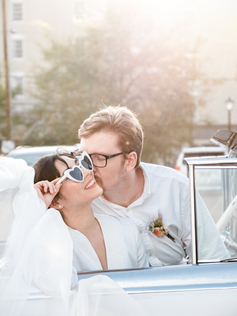 A woman wearing heart sunglasses laughs while her husband kisses her in a vintage car.