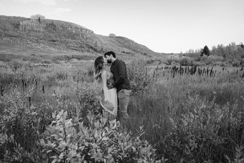Engaged couple kiss in a Alberta foothills field during their engagement session. He has his arms around her waist and she's holding her hat in her right hand