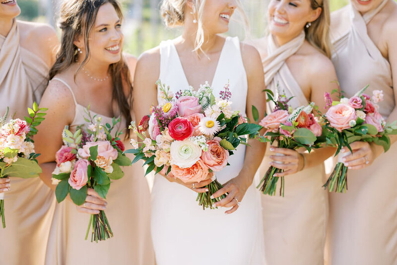 Bride with her bridesmaids in champagne color dresses holding colorful bouquets and smiling at each other