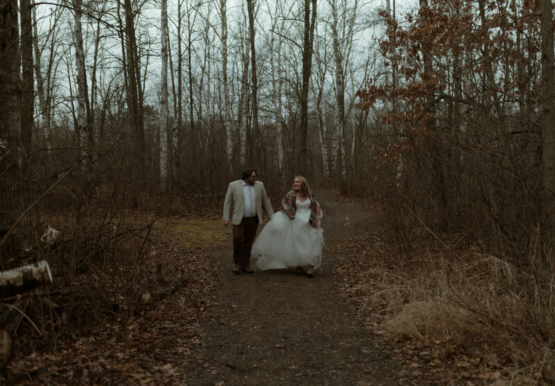couple walking hand-in-hand down a quiet forest path surrounded by tall, leafless trees on an overcast day. The bride wears a white gown with a cozy plaid shawl draped over her shoulders. The groom walks beside her in a beige blazer and dark trousers, both of them smiling and engaged in conversation as they stroll through the forest together.