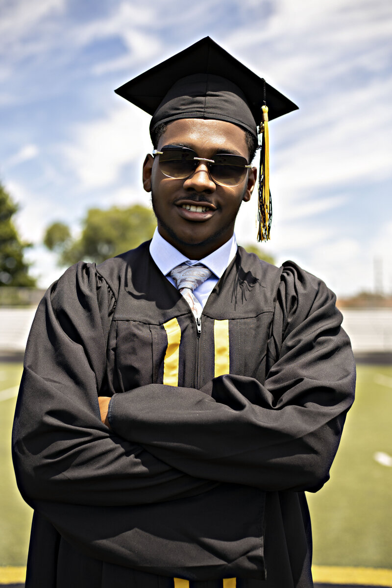 Senior boy with a light blue dress shirt, navy blue slacks, and sunglasses wearing a black graduation cap and gown on the football field