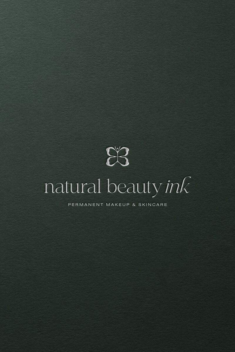 Debossed bespoke butterfly emblem logo for Natural Beauty Ink on green textured background.