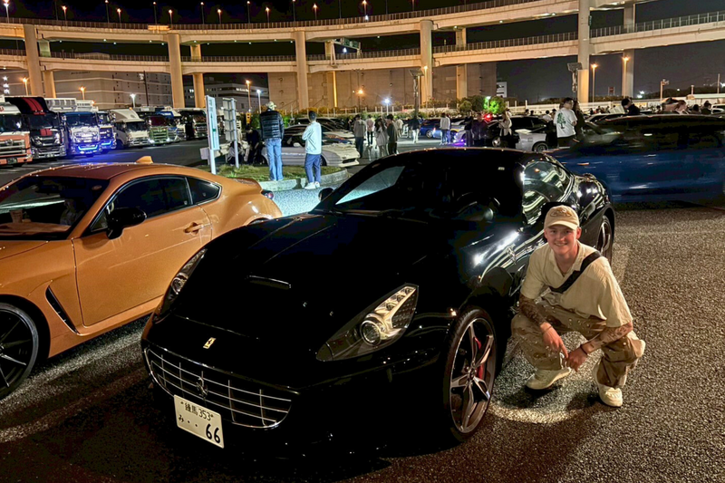 A black Ferrari parked under the glowing lights of Daikoku Parking Area at night, with a man crouching beside it and car enthusiasts gathered in the background.